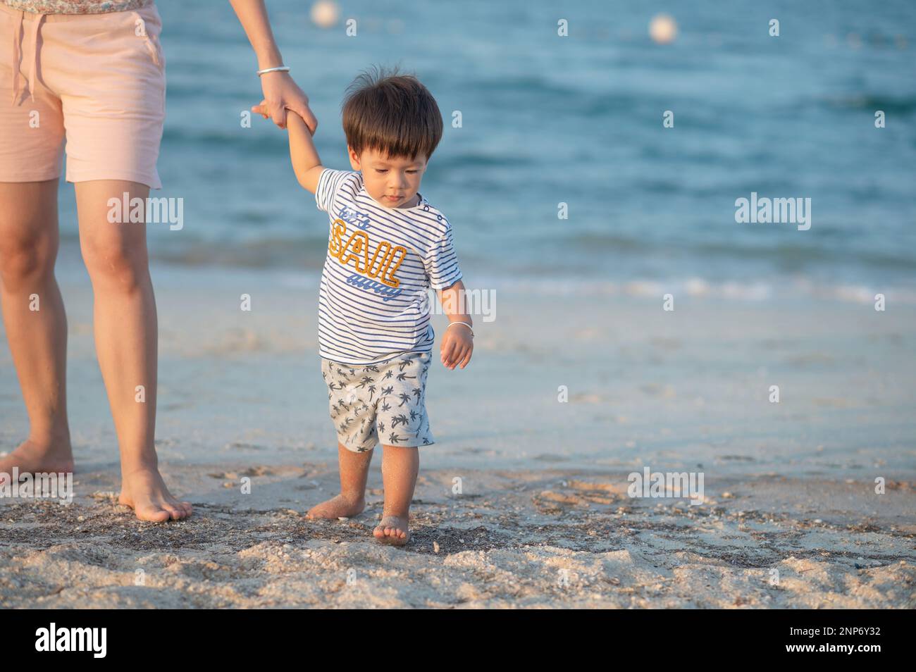 A mother and her toddler son, wearing casual clothing, walk hand-in-hand on a beach during a summer vacation Stock Photo