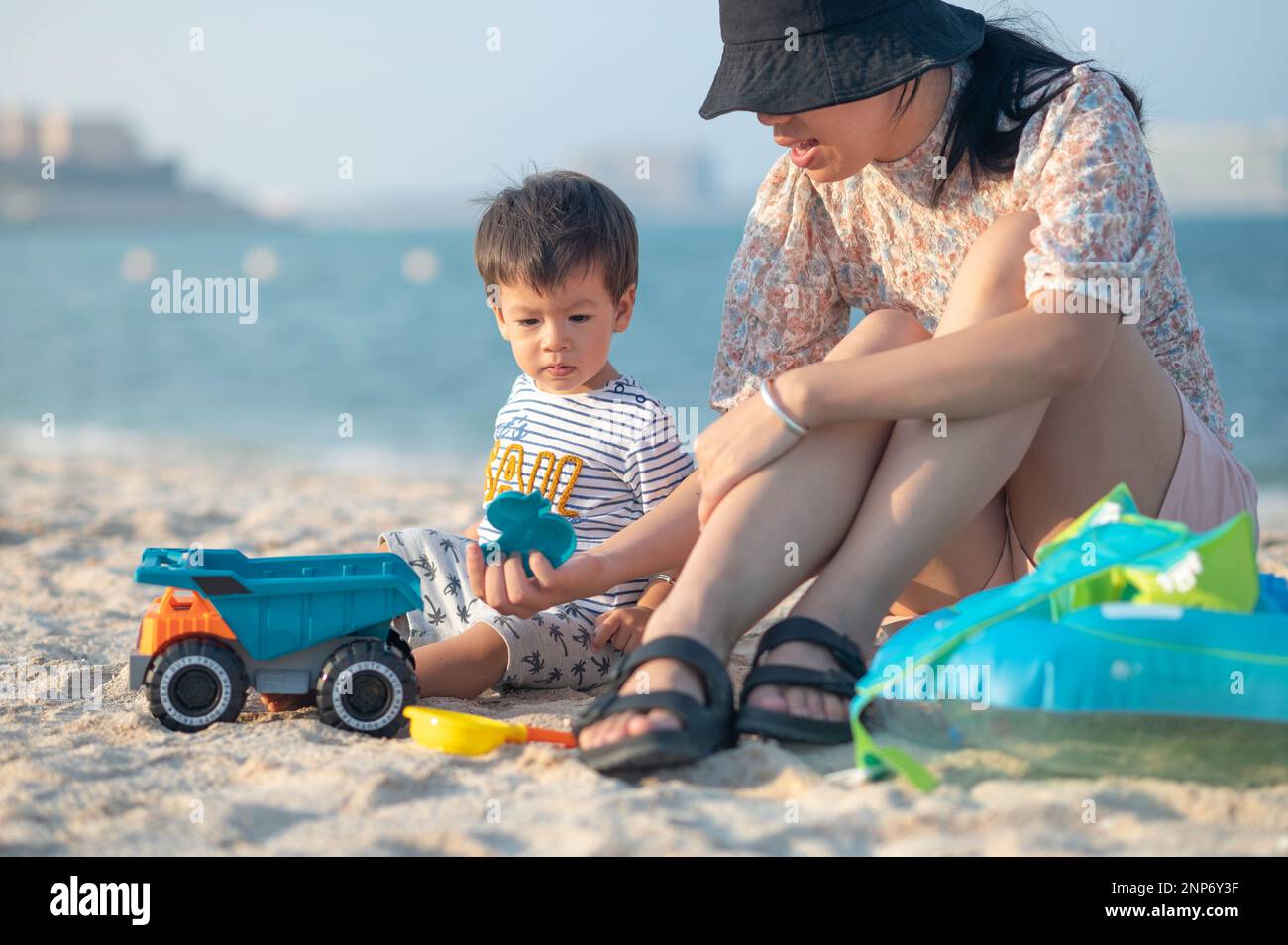 Mother playing with a toy truck and the sand on the beach with her baby boy enjoying the summer together by the seaside. Filled with joy and emotion t Stock Photo