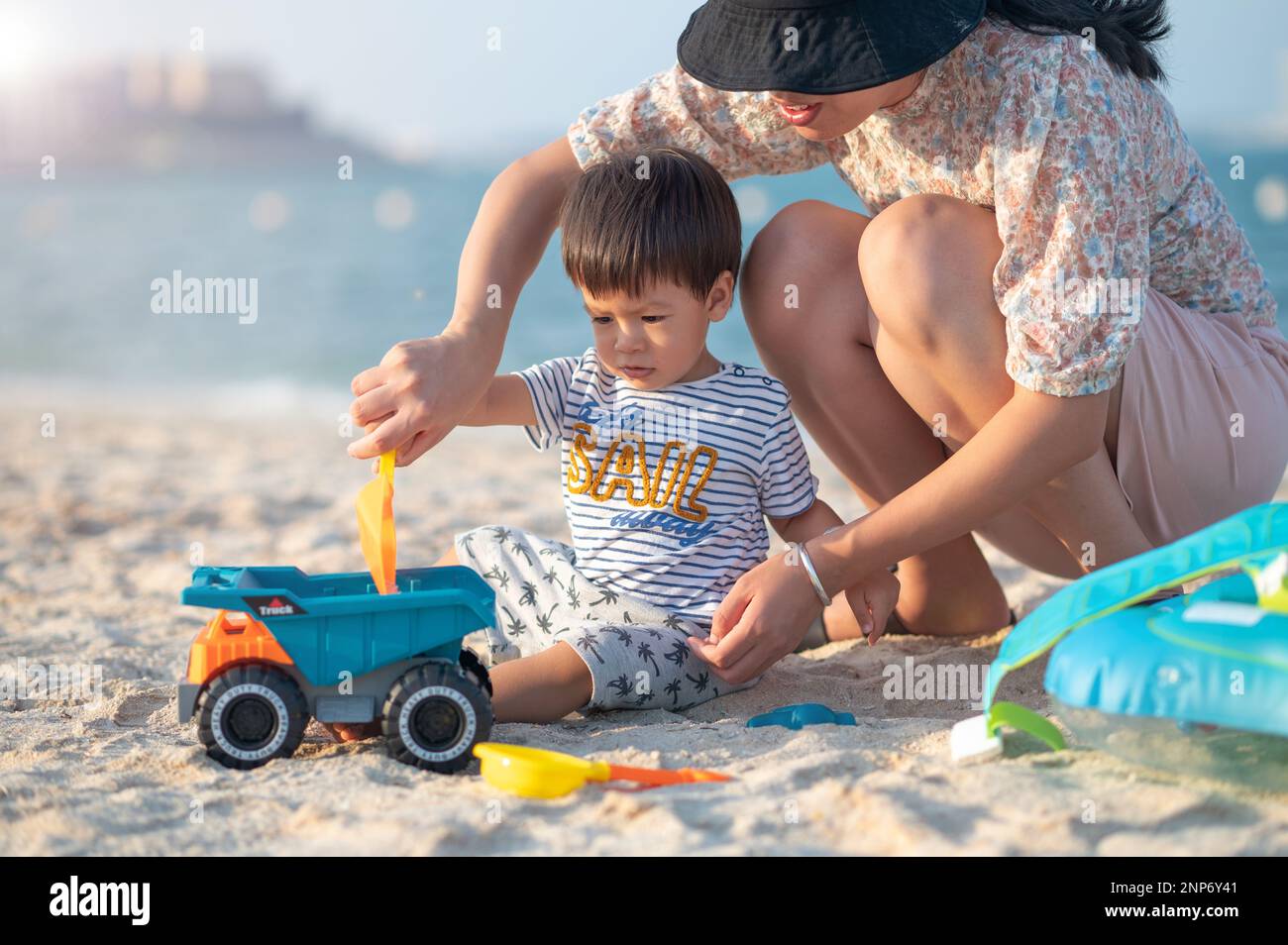 Mother playing with a toy truck and the sand on the beach with her baby boy enjoying the summer together by the seaside. Filled with joy and emotion t Stock Photo