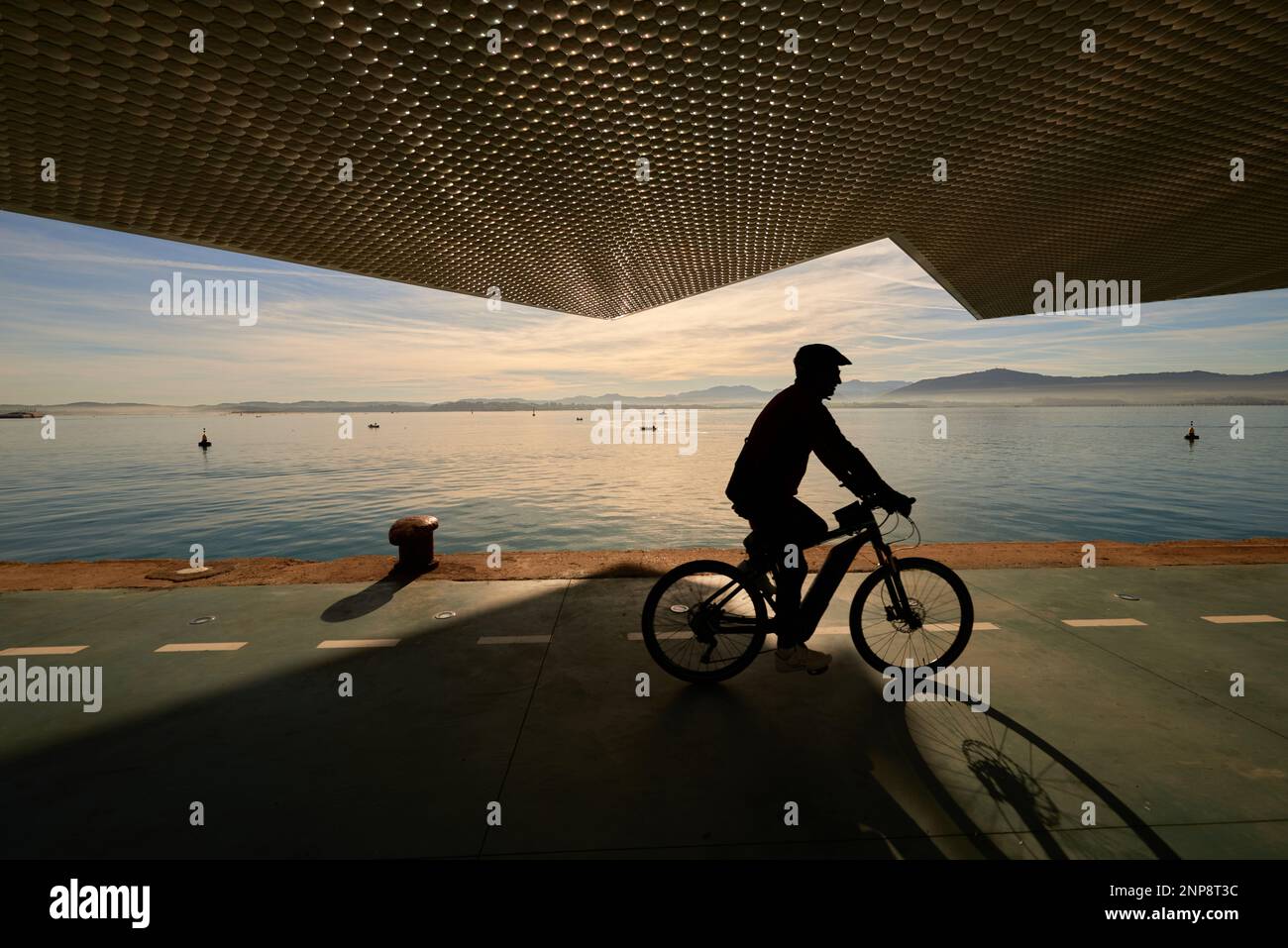 Santander Bay seen from the Botín Center while a cyclist against the light circulates with an electric bicycle along the pier with the sky cut out by Stock Photo