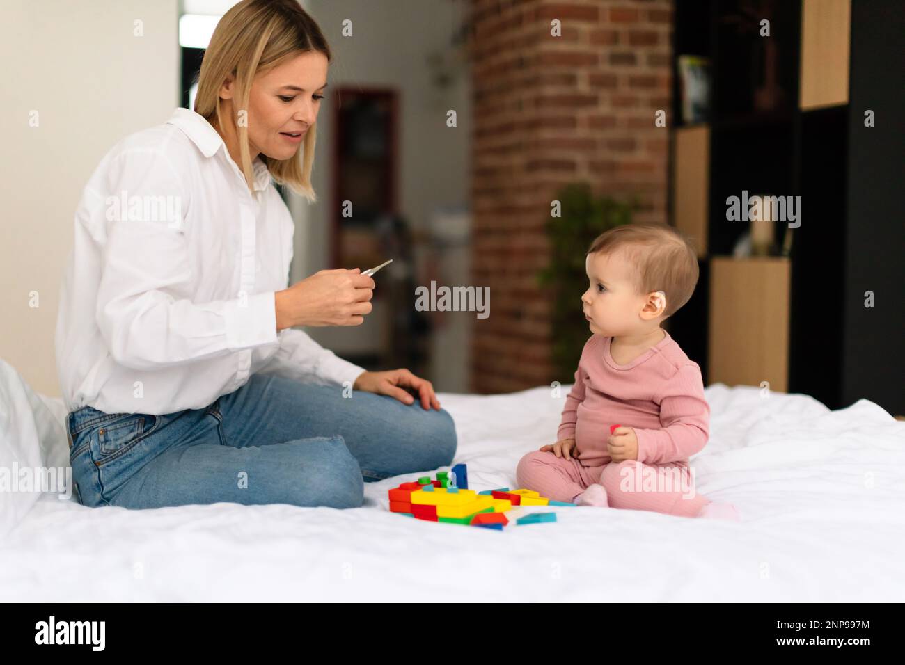 Mom measuring temperature to her daughter child with thermometer, sitting on bed in bedroom, free space Stock Photo