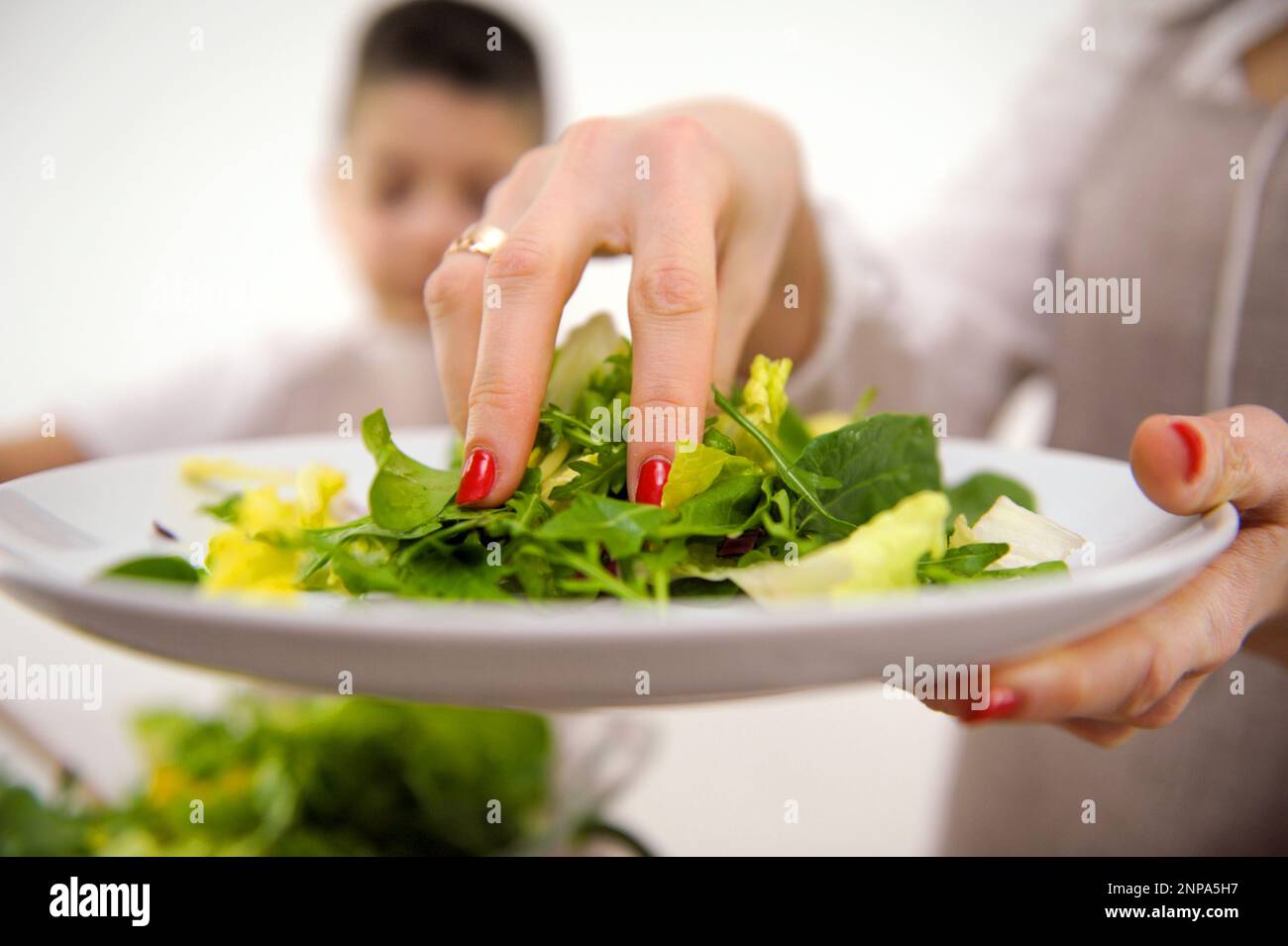 cooking salad mom with hand adds lettuce leaves to large bowl in background boy stirs salad kitchen cooking aprons cooking homemade food communication children helping child in cooking training Stock Photo