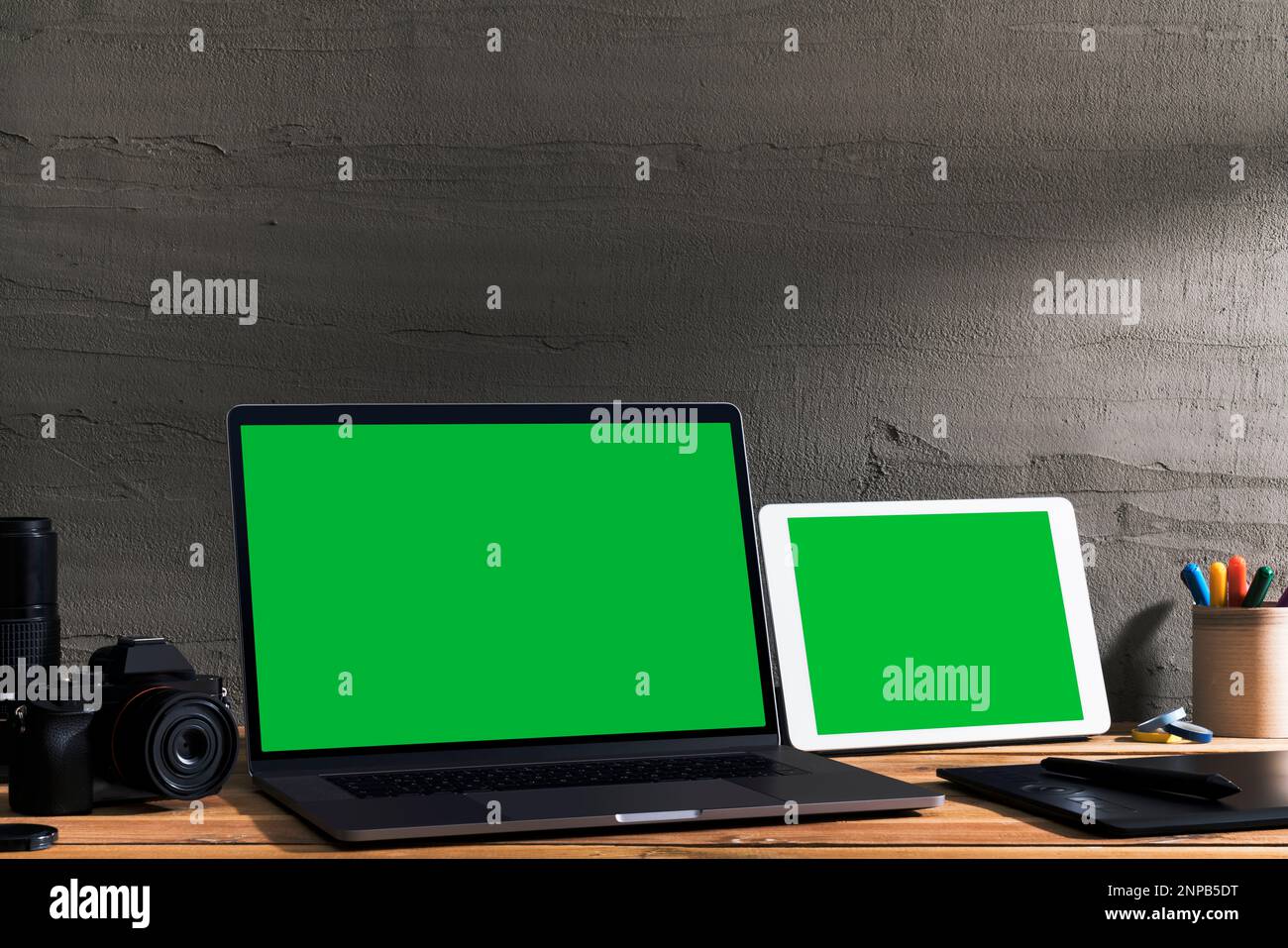Chroma key green screen, angled view laptop and digital tablet on table with digital photography equipment on wooden table in front of concrete wall. Stock Photo