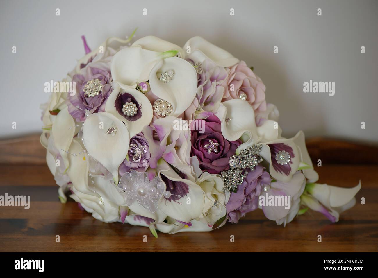 Cascading wedding bouquet with calla lilies, purple roses, decorated with silver jewelry, silk and pearl beads rested on a wooden table Stock Photo