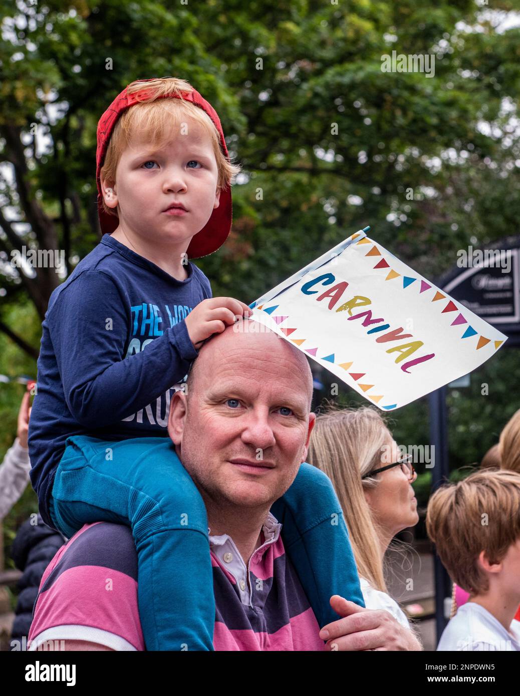 A boy sits on his dad's shoulders with a Carnival flag and looks on at