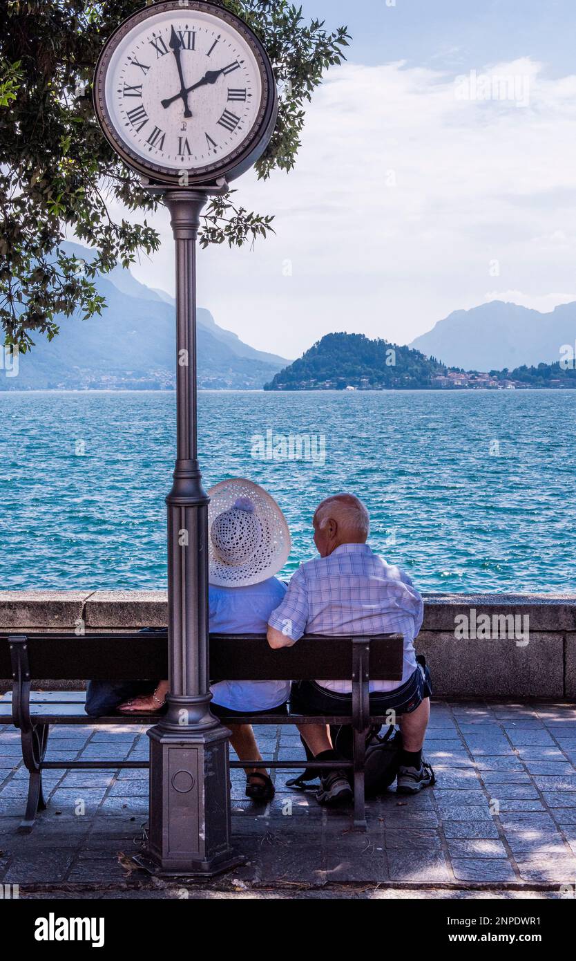 An old couple looks out at Lake Como from a bench in Menaggio with a street clock just behind them. Stock Photo