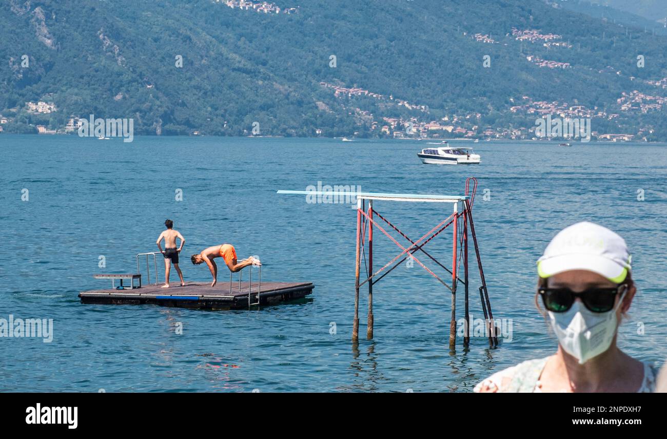 A man is caught in midair as he is jumping in the water on Lake Como while a woman wearing sunglasses and a face mask enters the frame in the foregrou Stock Photo