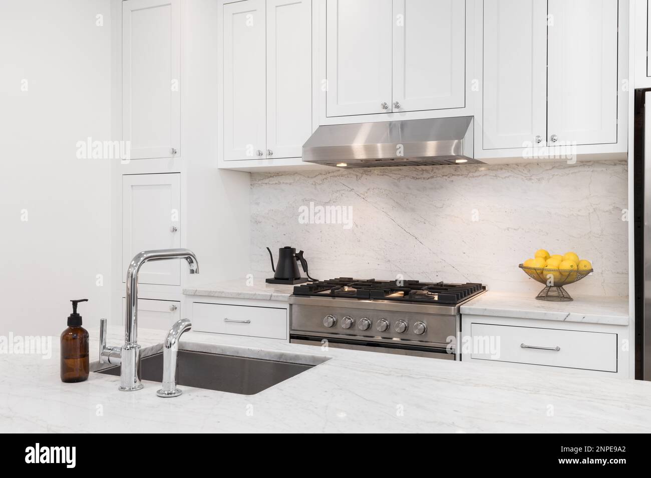 A kitchen detail with white cabinets, stainless steel stove and hood, marble countertops and backsplash, and a chrome sink. Stock Photo