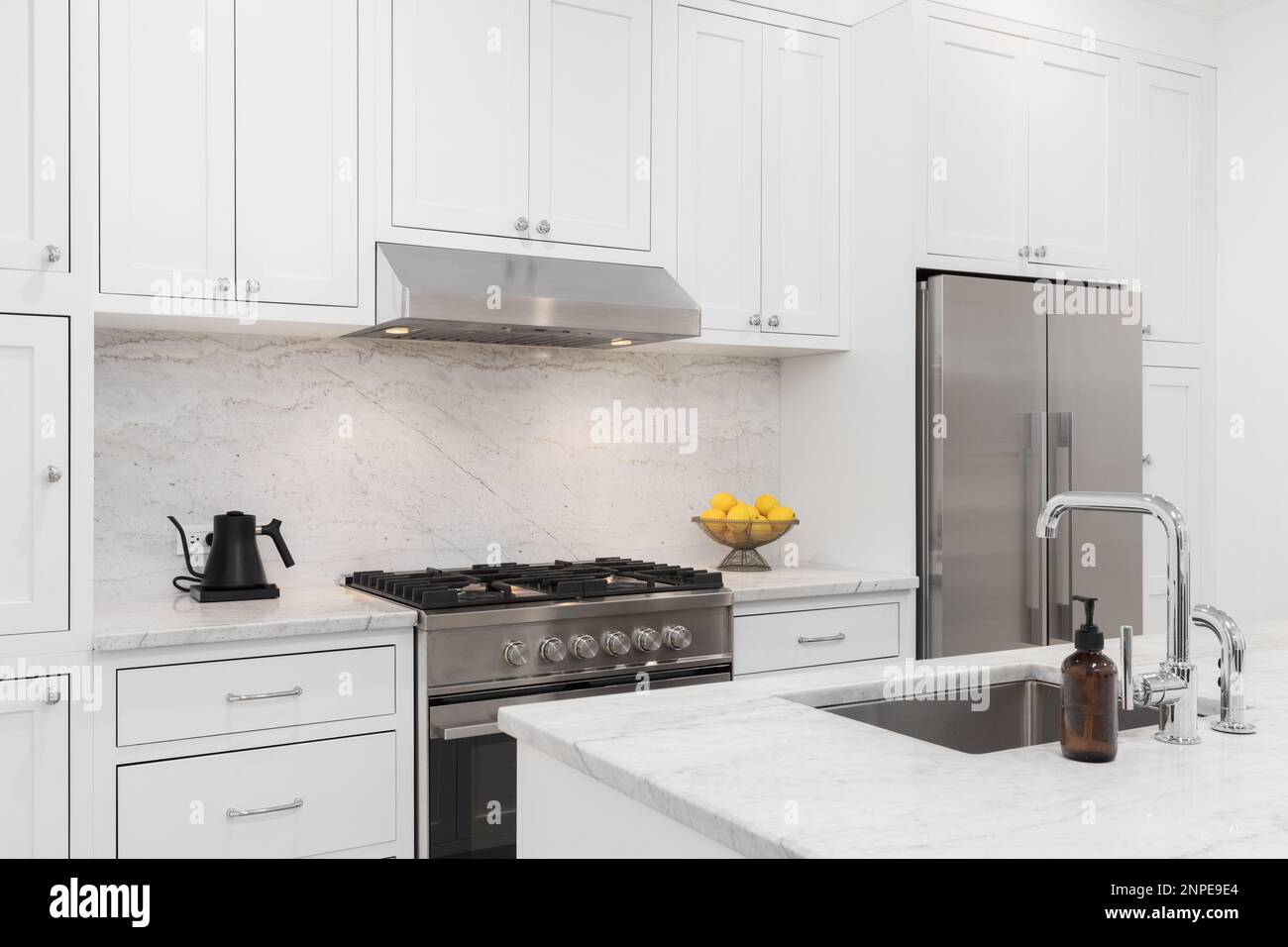 A kitchen detail with white cabinets, stainless steel stove and hood, marble countertops and backsplash, and a chrome sink. Stock Photo
