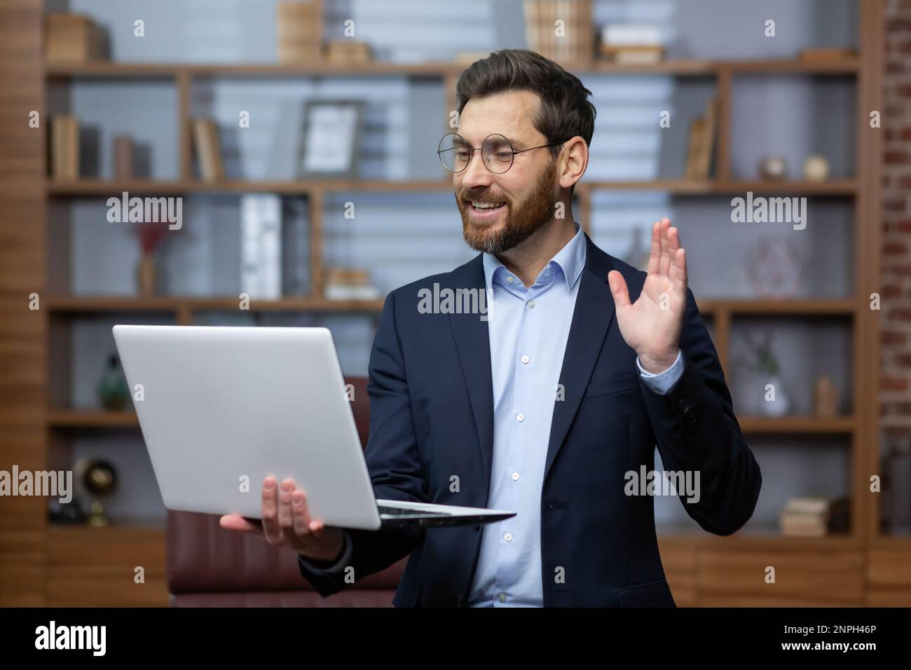 A mature male lawyer inside the office is standing at the desk, the businessman boss is talking remotely via video call using a laptop, man chatting with colleagues, advising online clients. Stock Photo