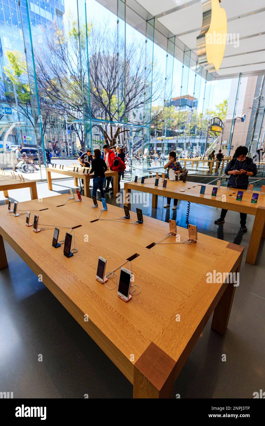 Interior of the Apple store in Omotesando, Tokyo. People looking at the latest I-phones displayed on long wooden tables in glass building. Daytime. Stock Photo