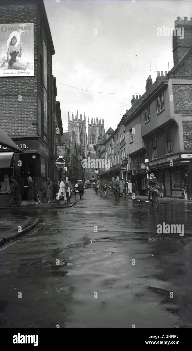 1950s, historical, distance view of York Minster from Low Petergate, York, England, UK. Founded in 627 AD, the Anglican cathedral is one of the world's most spectacular. A street in the city centre, Petergate is divided into High Petergate and Low Petergate. On the left, on a wall of a building is a billboard or poster of a young lady in a swmming costume and sun hat, with the line 'Ovaltine is Delicious & Refreshing'.  Also in the picture are signs for; Petergate Fish Restaurant, Merrimans, The Kettering & Leicester Boot Co & Surgical Stores. Stock Photo
