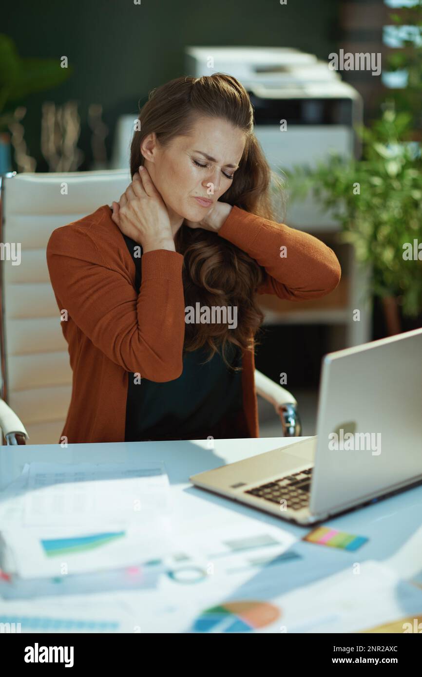 sad trendy 40 years old small business owner woman with laptop and neck pain in the modern green office. Stock Photo
