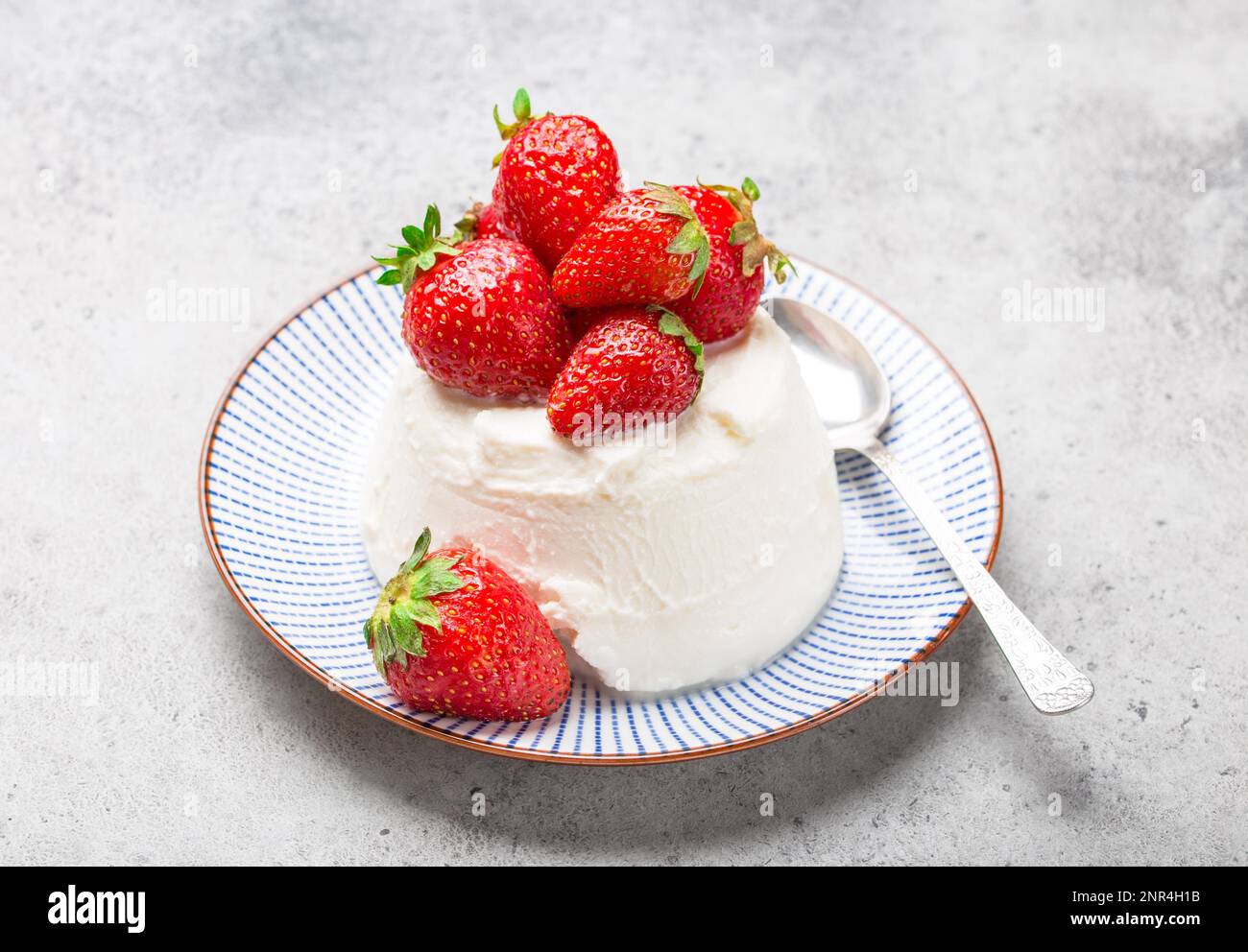 Fresh Italian cheese ricotta with strawberries on a plate with a spoon, grey rustic stone background, light summer dessert or snack, good for diet or Stock Photo