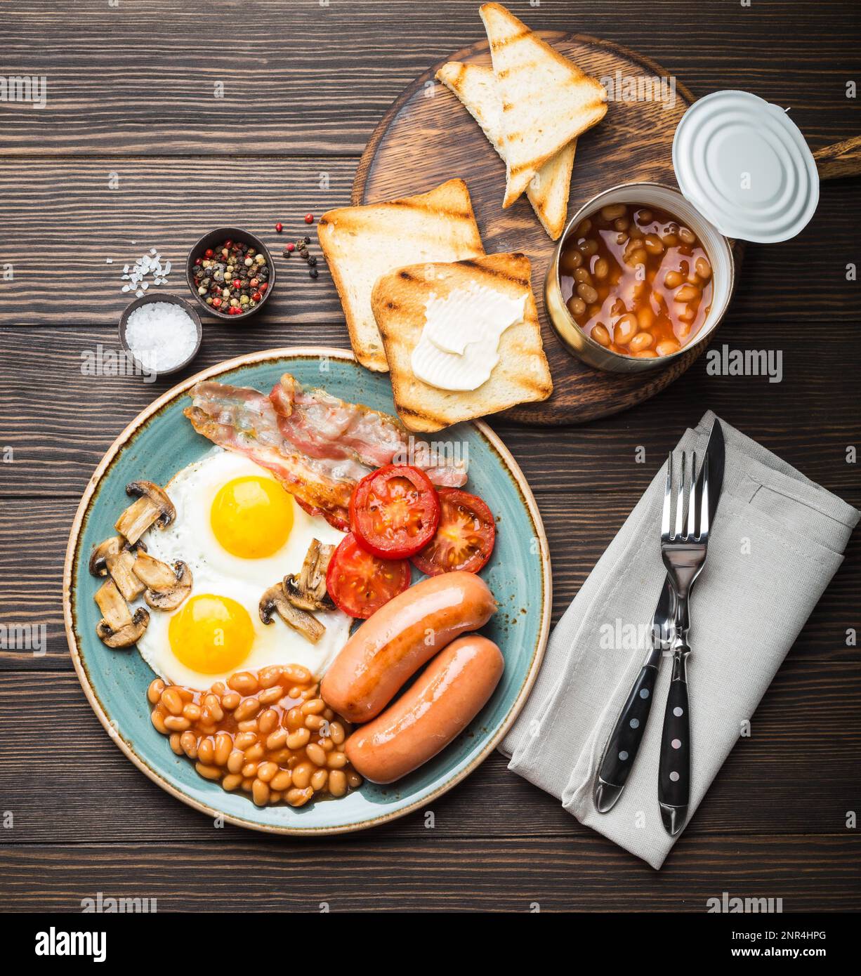 Full English breakfast with fried eggs, sausages, bacon, beans, mushrooms, tomatoes on a plate, bread toasts with butter. Traditional British meal Stock Photo