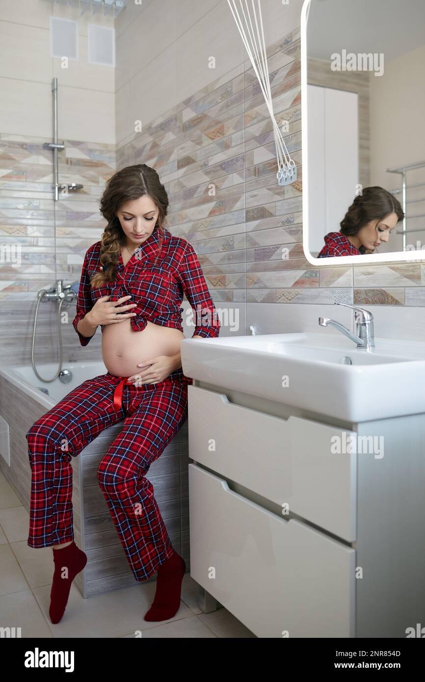 A pregnant woman in the bathroom sitting on the edge of the bath. Female dressed in red pajama ...
