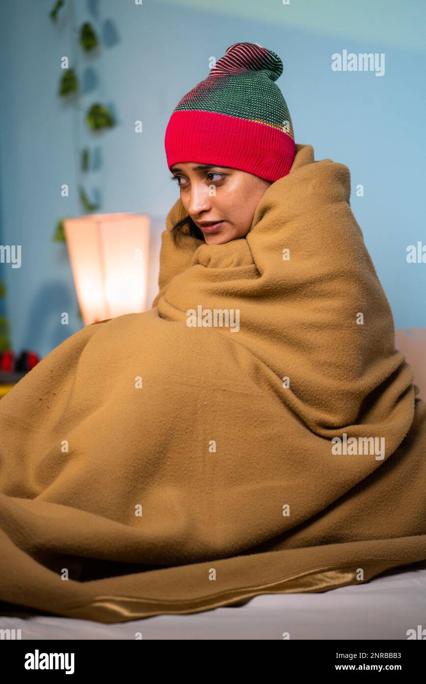 Vertical shot of shivering Young girl full coverd with blanket and cap sitting on bed at during night - concept of chilling winter cold, climate Stock Photo