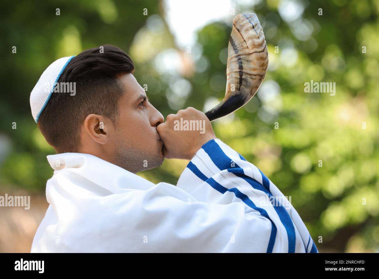 Jewish man in kippah and tallit blowing shofar outdoors. Rosh Hashanah
