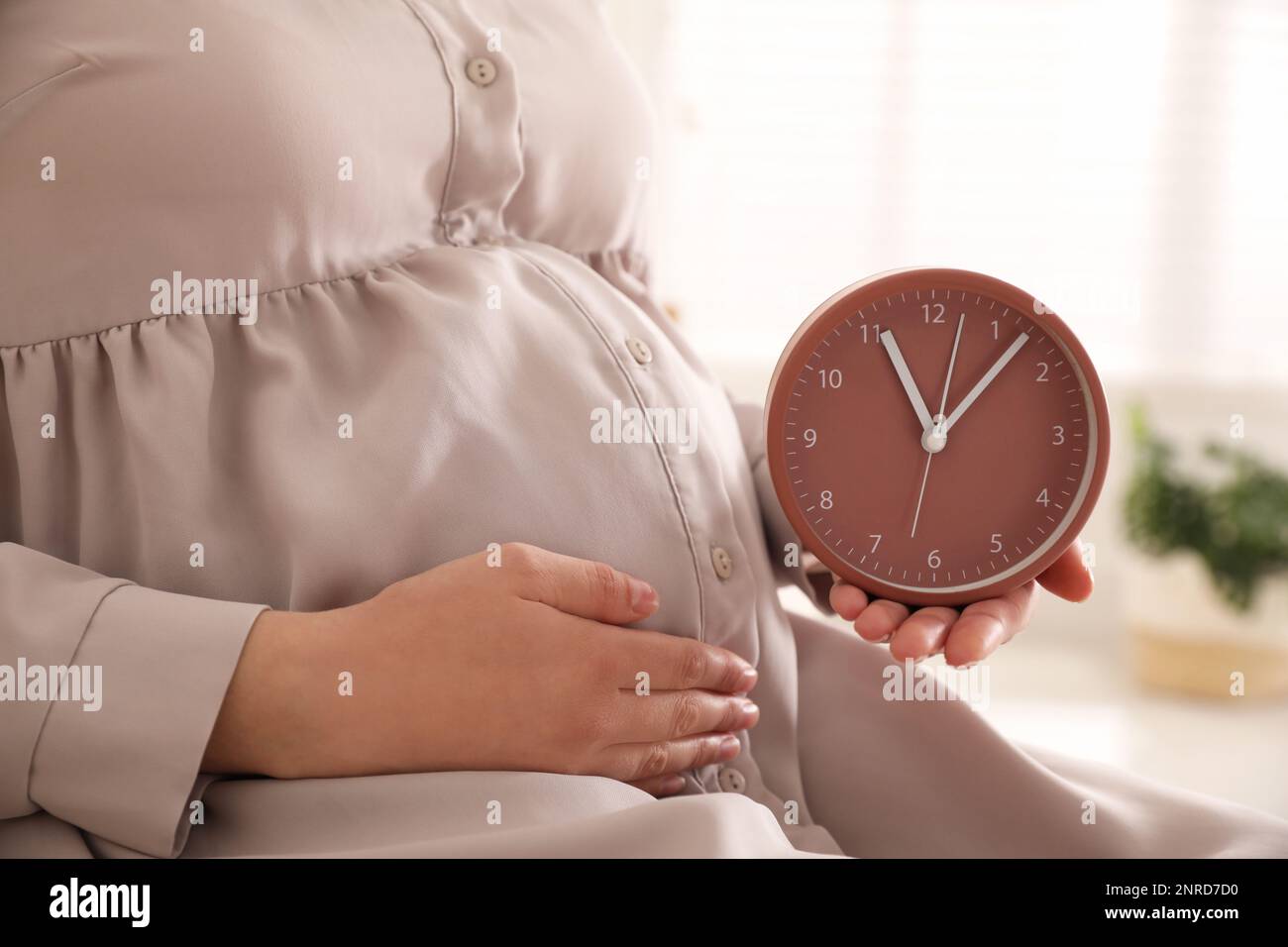 Young pregnant woman holding clock near her belly at home, closeup. Time to give birth Stock Photo
