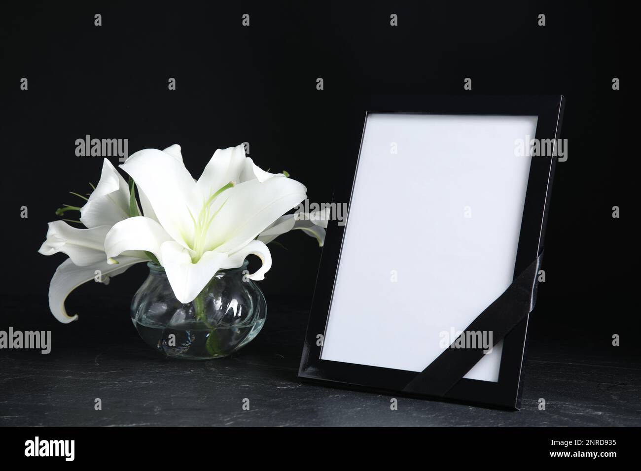 Funeral photo frame with ribbon and white lilies on black table against dark background. Space