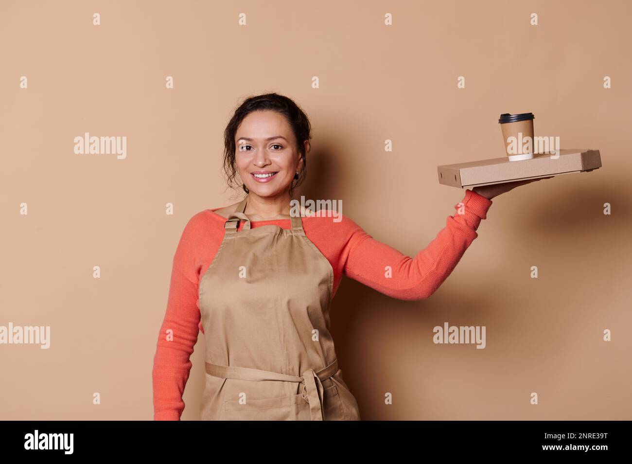 Stunning female bartender barkeeper barista waitress smiles at camera, holds flatbox and eco paper cup of takeaway drink Stock Photo