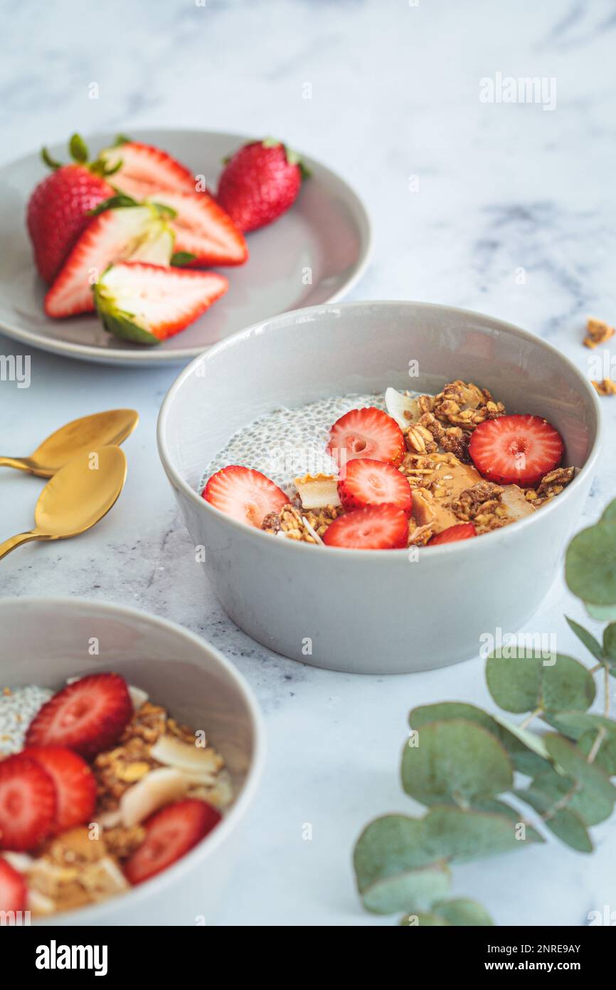 Chia pudding with homemade coconut granola, peanut butter and strawberries in gray bowl, marble background. Healthy plant based diet, detox, summer re Stock Photo
