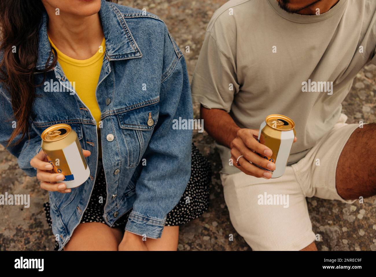 Midsection of male and female friends holding drink cans while sitting together on rock Stock Photo
