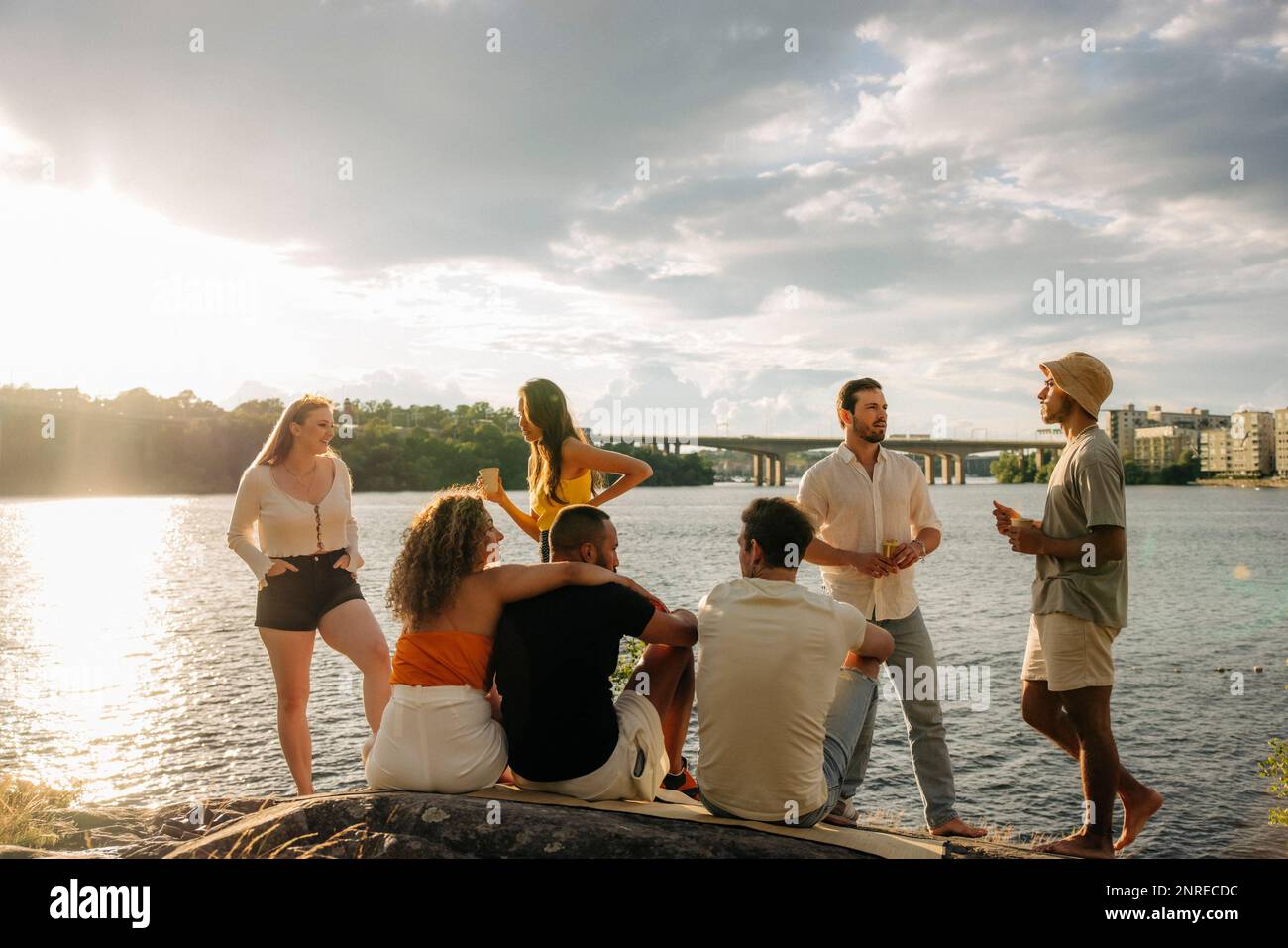 Multiracial male and female friends talking with each other during picnic near sea at sunset Stock Photo