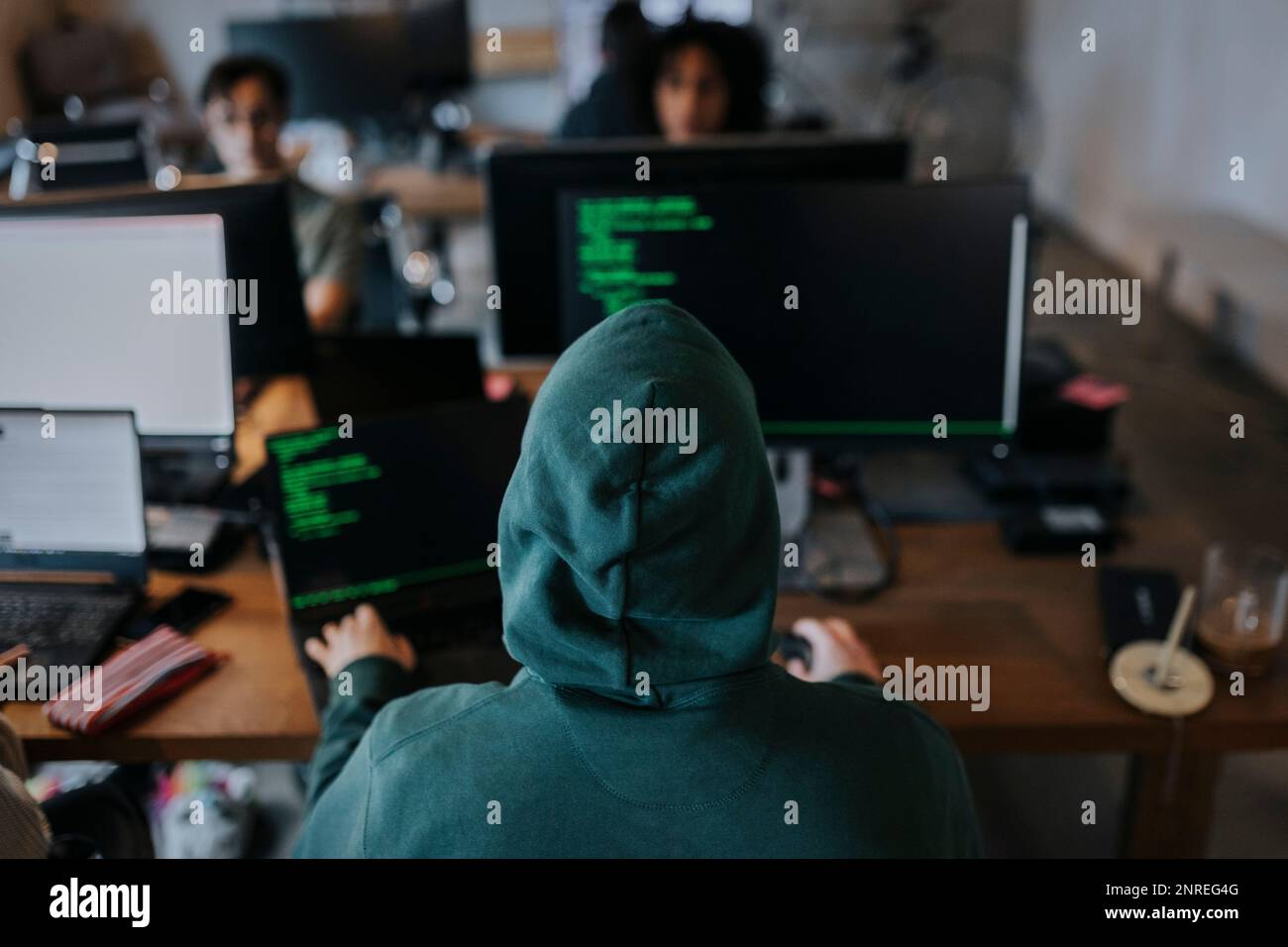 Rear view of young male hacker wearing hood coding on computer at desk in creative workplace Stock Photo