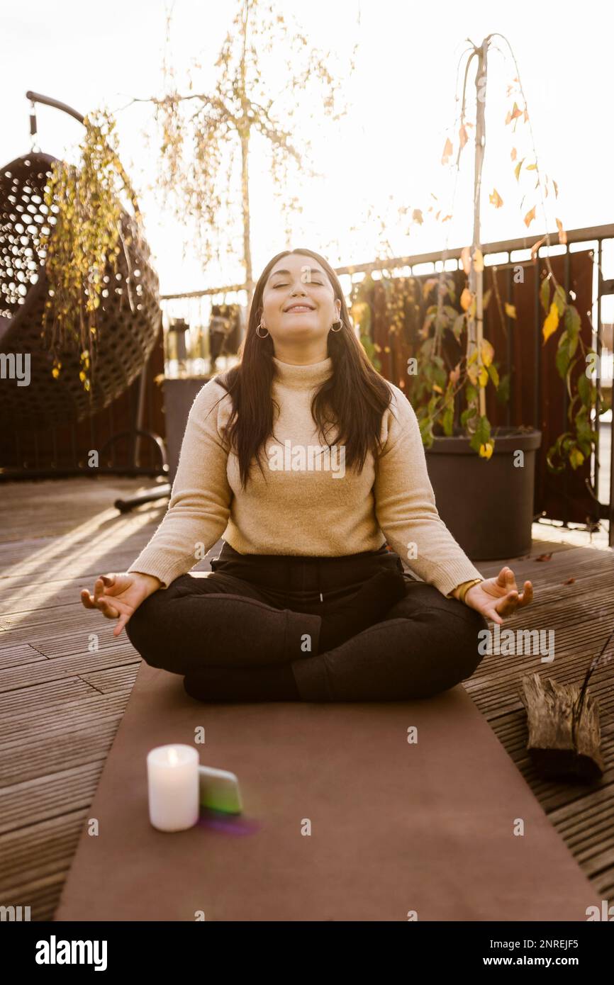 Smiling woman practicing Lotus position on exercise mat at terrace Stock Photo