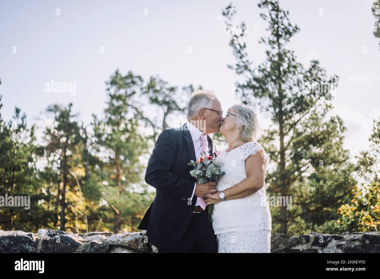Affectionate newlywed senior couple kissing on mouth against forest on wedding day Stock Photo