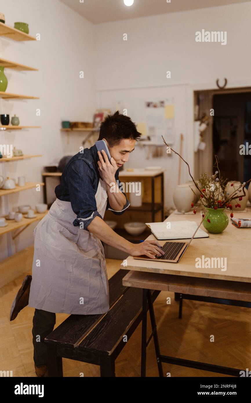 Potter talking on smart phone while using laptop on table at workshop Stock Photo