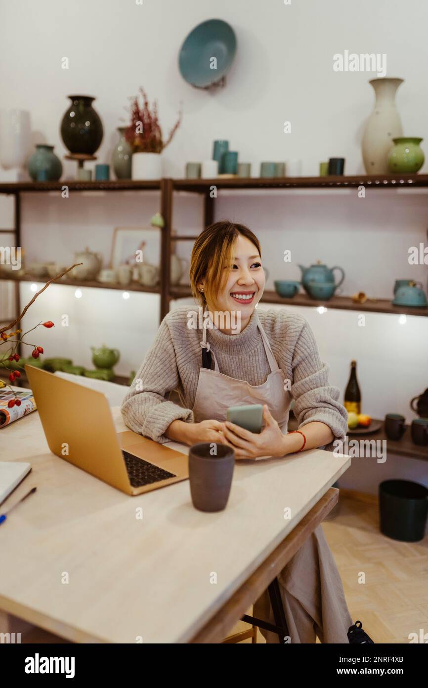 Happy female potter with laptop and smart phone sitting at workshop Stock Photo