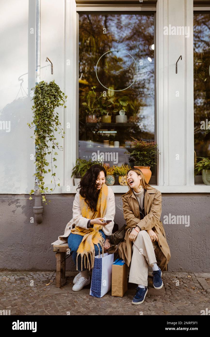 Happy female friends talking to each other while sitting on bench outside store Stock Photo