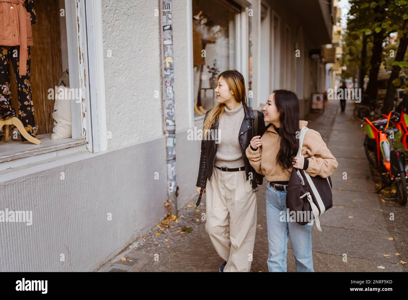 Female friends looking at store window while walking on sidewalk Stock Photo