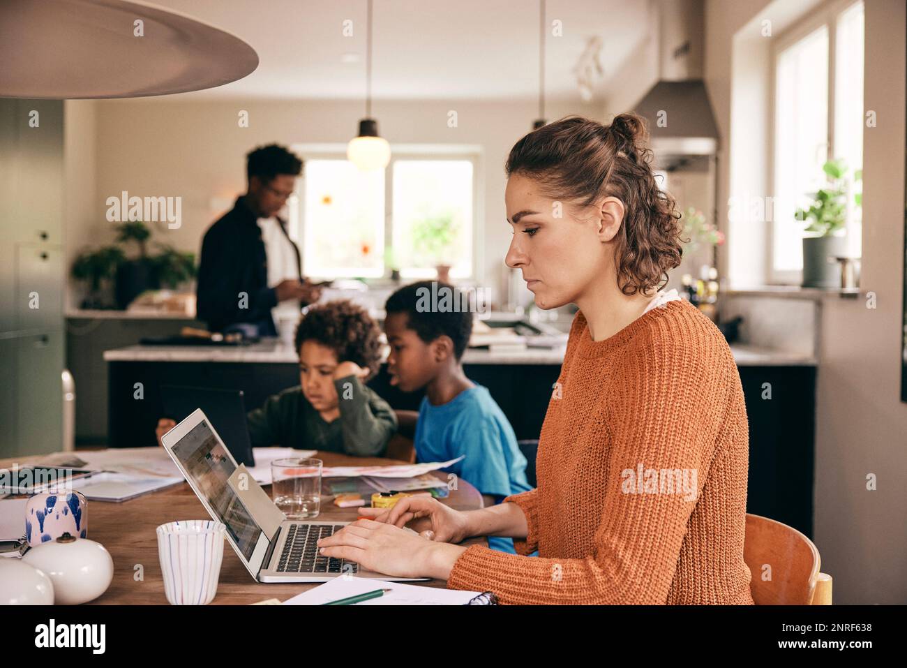 Side view of woman using laptop while family in background at home Stock Photo