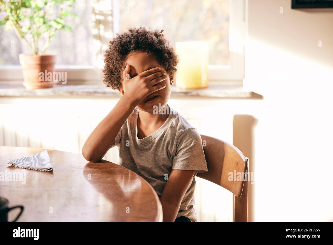 Multiracial boy covering eyes with hands while sitting on chair at home Stock Photo