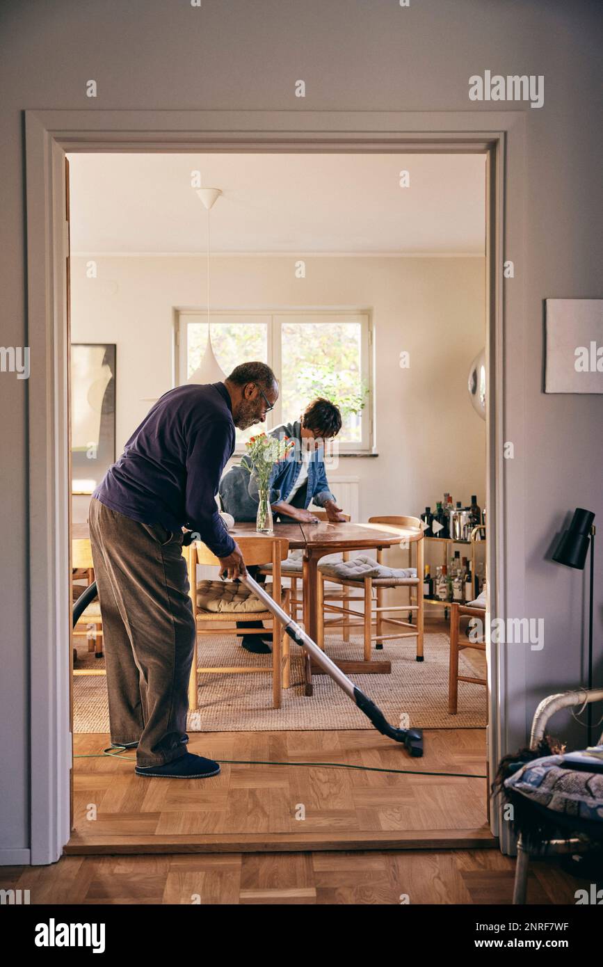 Side view of senior man cleaning floor with vacuum cleaner seen through doorway at home Stock Photo