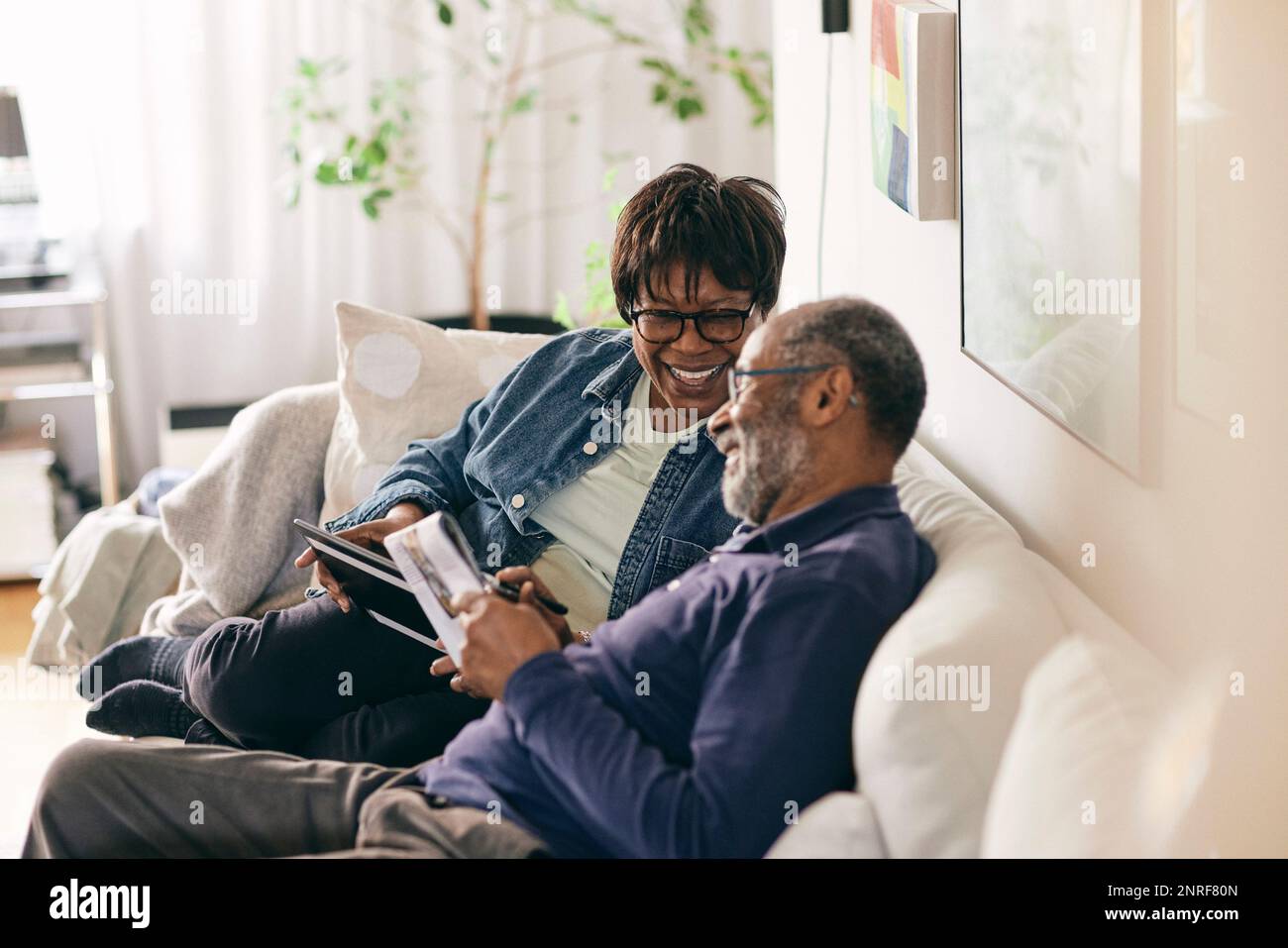 Happy senior woman with digital tablet talking to man sitting on sofa at home Stock Photo