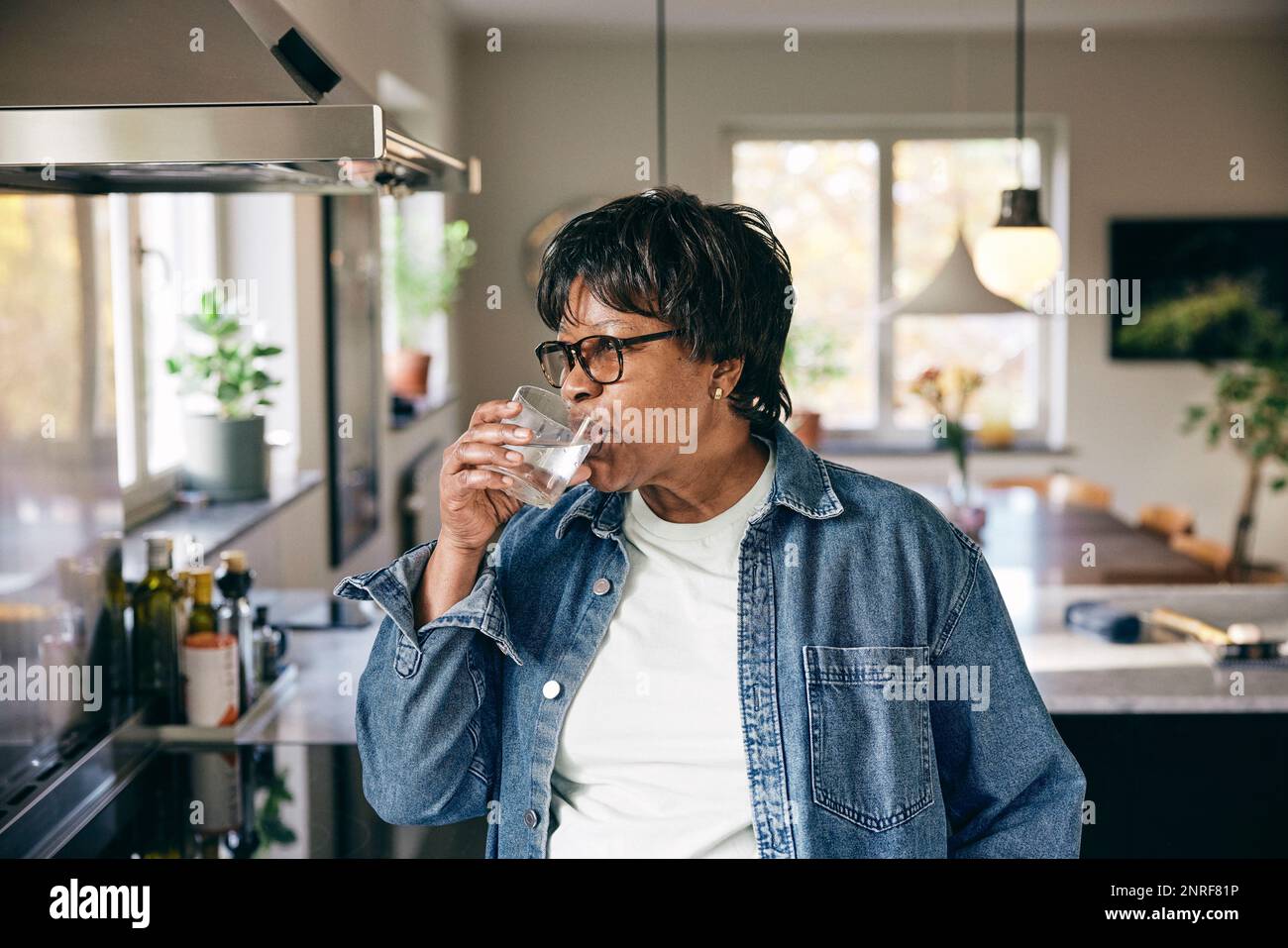 Senior woman wearing denim jacket drinking water in kitchen at home Stock Photo