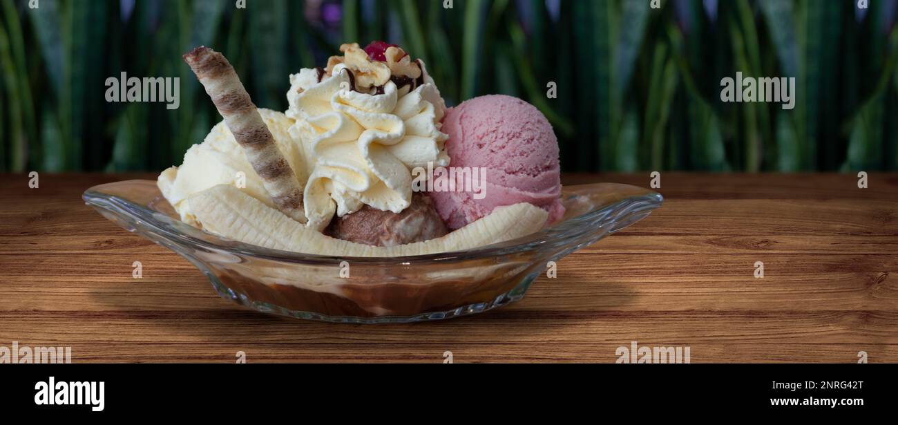 Banana split garnished with whipped cream and chocolate sorbet in clear glass duck on wooden table with defocused green plants background. Image with Stock Photo