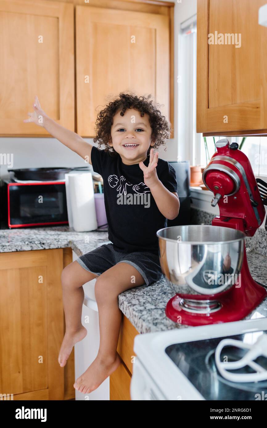 Excited celebrating biracial toddler baking and sitting on counter Stock Photo