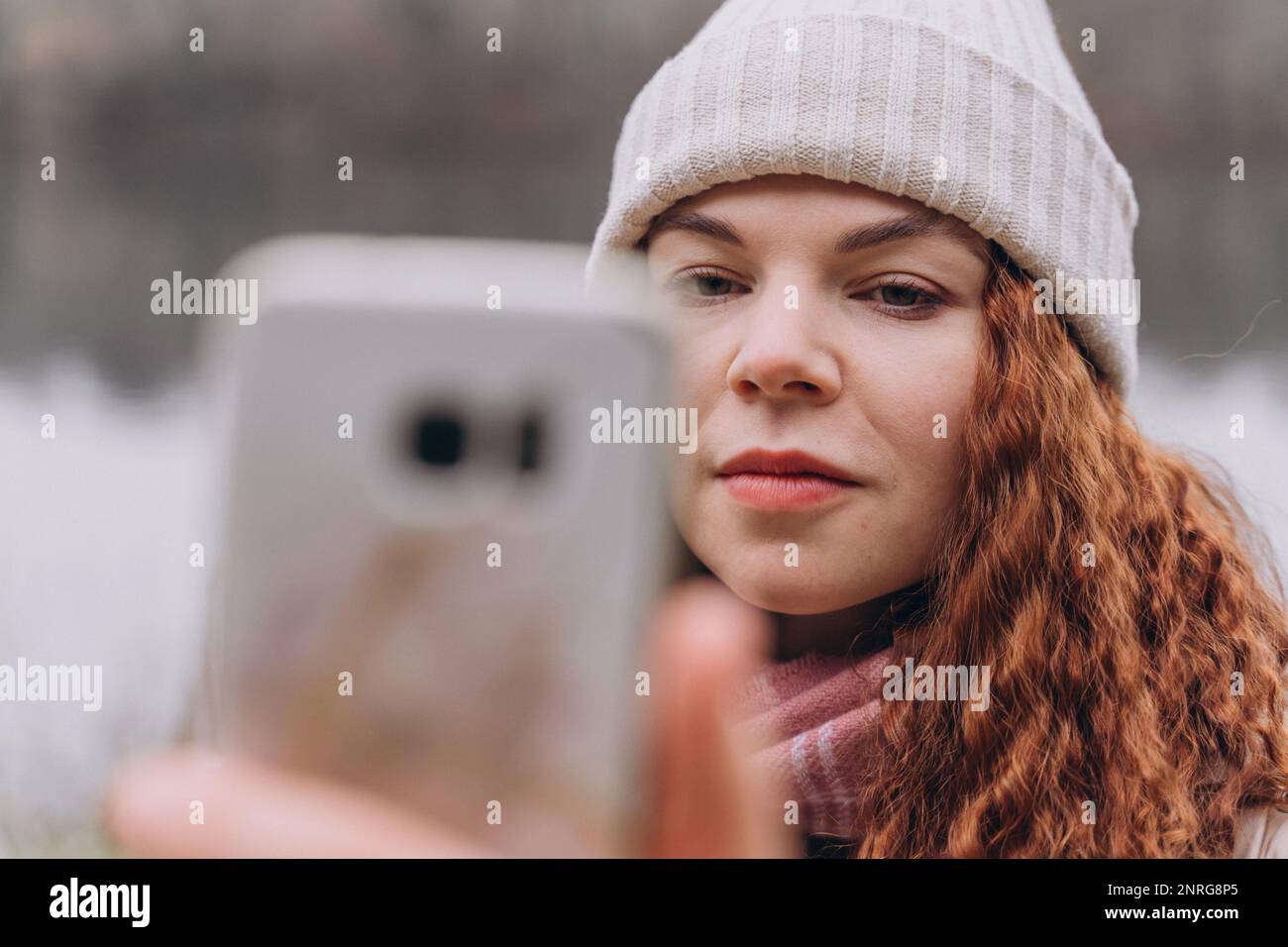 Curly woman 30-35 in a knitted hat looks seriously her phone Stock Photo