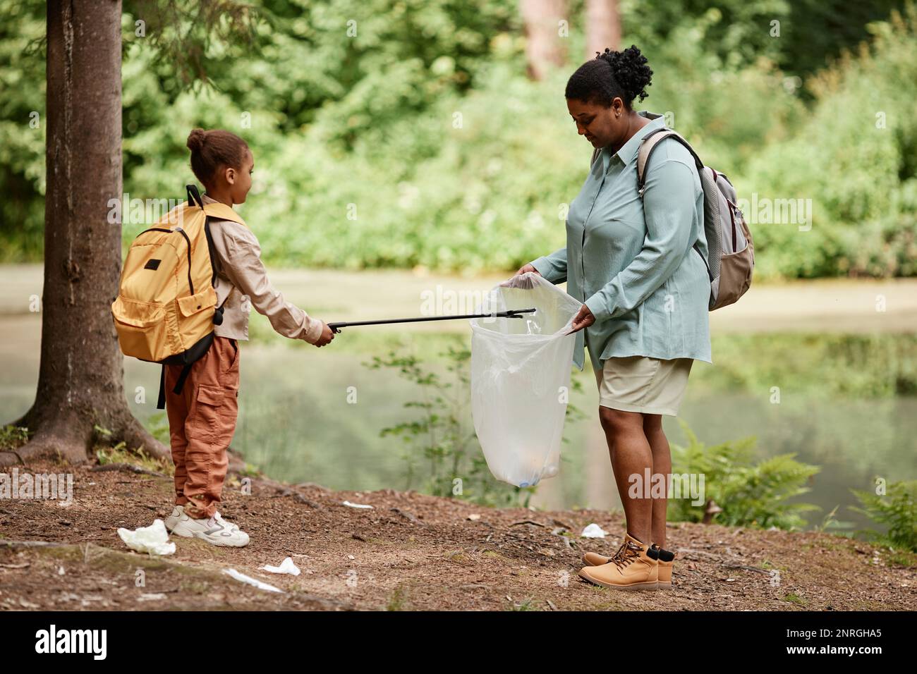 Side view portrait of black mother and daughter helping cleaning nature together and picking up trash by river, copy space Stock Photo