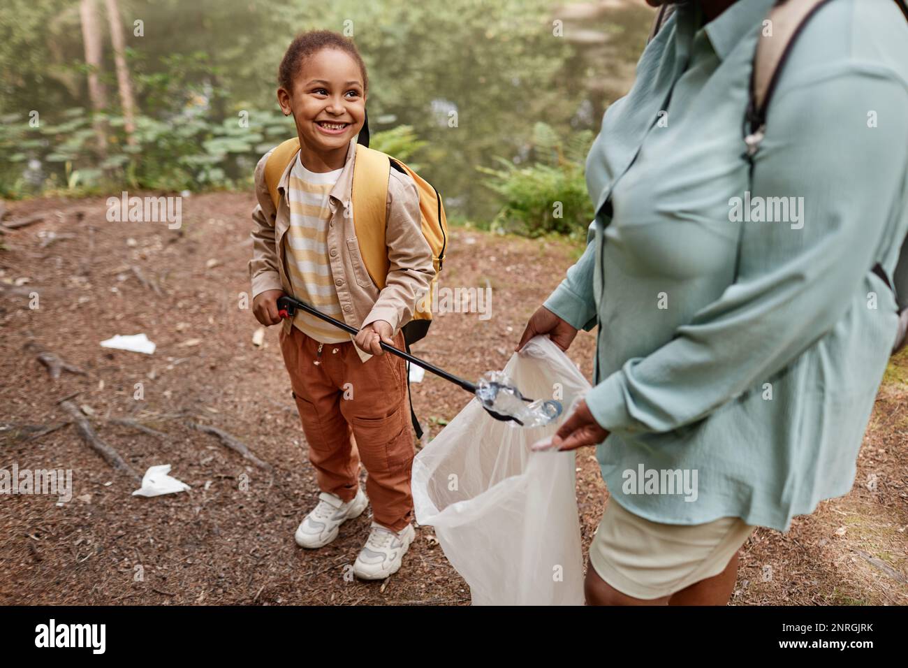 High angle portrait of smiling black girl helping clean nature and picking up plastic bottles outdoors Stock Photo