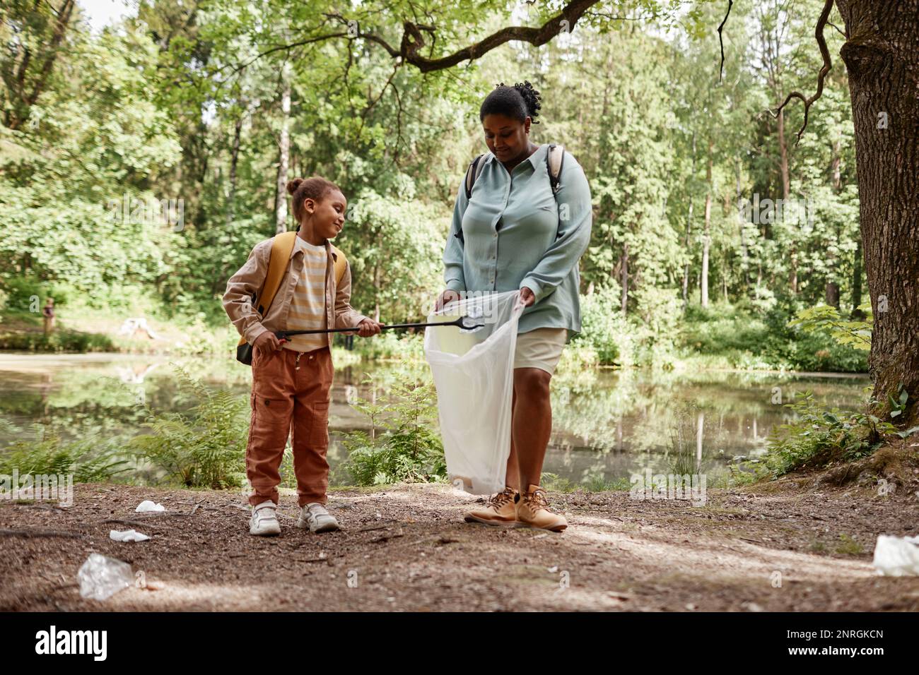 Full length portrait of black mother and daughter helping clean nature and picking plastic bottles in forest Stock Photo