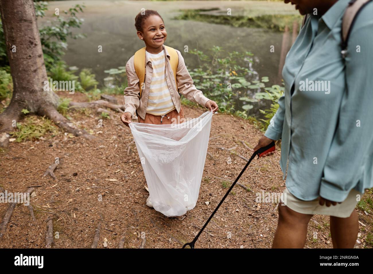 High angle portrait of smiling black girl holding trash bag outdoors and helping clean nature with mom Stock Photo