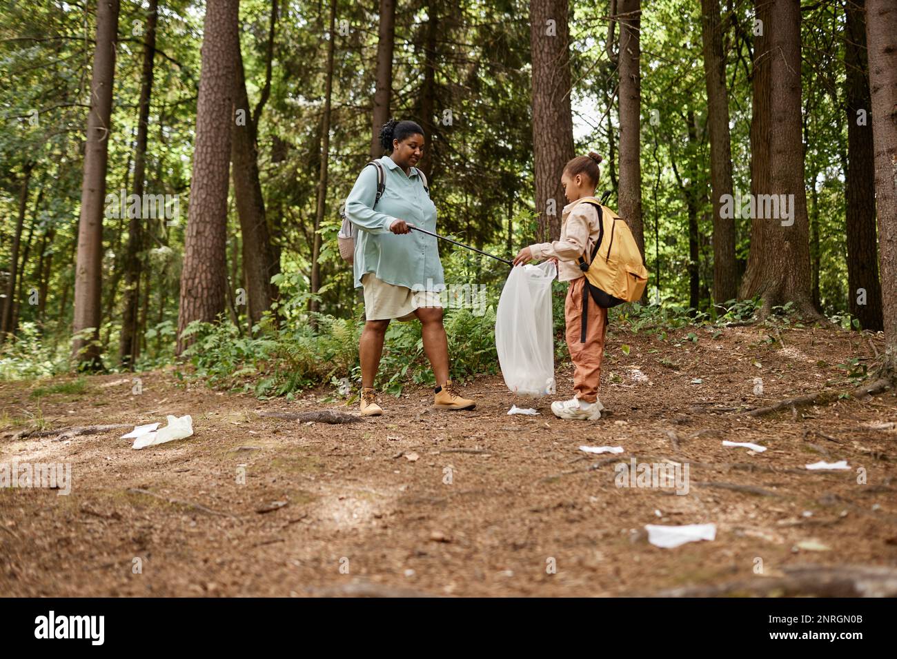 Side view portrait of mother and daughter picking plastic bottles on nature trail, copy space Stock Photo