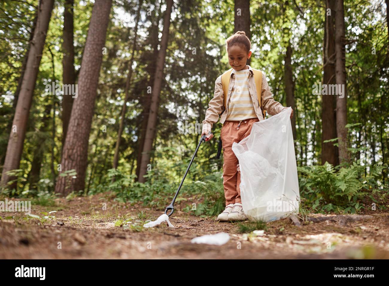Full length portrait of smiling little girl picking up trash in nature trail, copy space Stock Photo