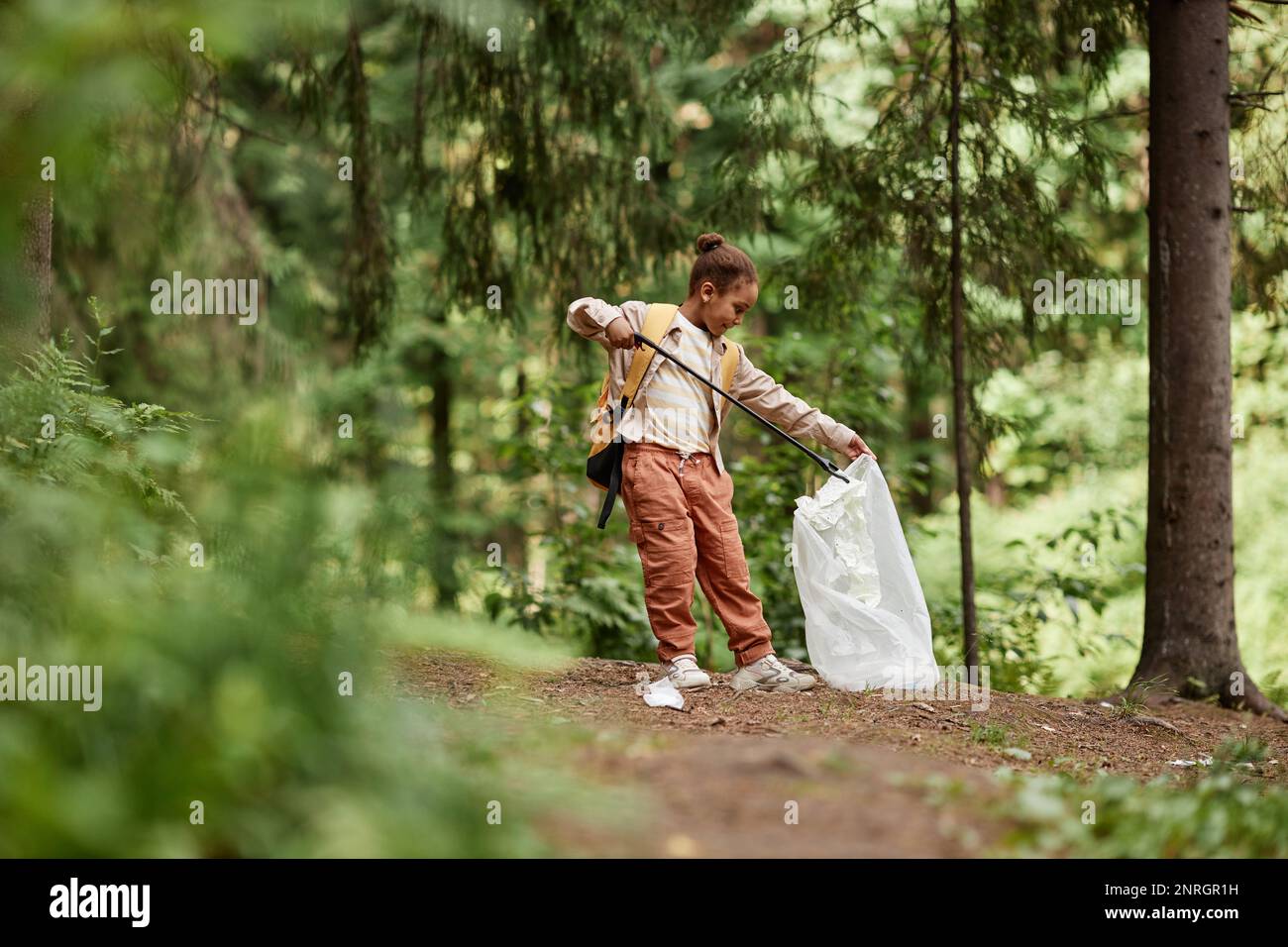 Full length portrait of black little girl picking up plastic bottles in nature trail, copy space Stock Photo